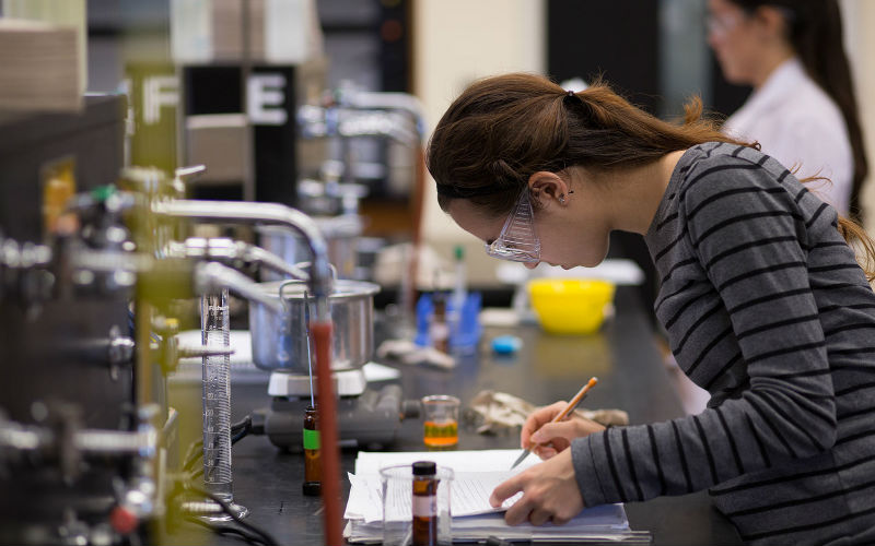 A student takes notes while working in a lab with chemistry equipment